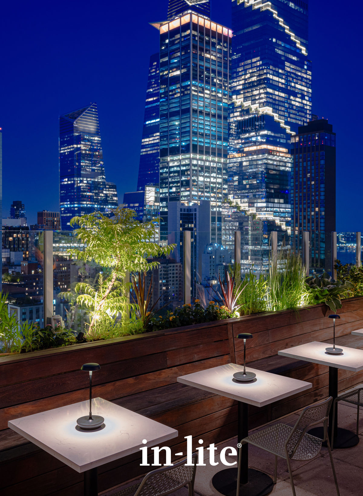in-lite outdoor lighting illuminating a rooftop hospitality terrace with tables, planters, and a city skyline at night